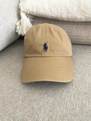 Beige keps från Polo Ralph Lauren unisex - Snygg beige keps från Polo Ralph Lauren med klassisk broderad logga i mörkblått framtill och 'POLO' broderat baktill. Justerbar rem med metallspänne. Perfekt accessoar för en avslappnad och stilren look.