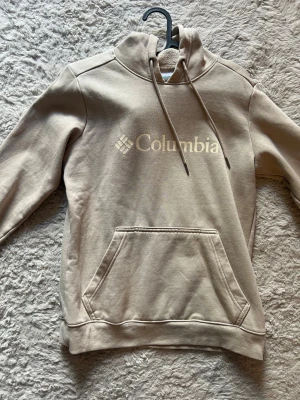 Beige hoodie från Columbia - Snygg beige hoodie från Columbia med stor logga på bröstet och mindre logga på ärmen. Klassisk känguruficka framtill och justerbar huva med dragsko. Tillverkad i mjukt och bekvämt material, perfekt för en avslappnad stil.