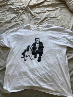 Ulf Kristersson tröja - Säljer en vit t-shirt från Fruit of the Loom med ett svart tryck av en man i kostym och en hund på framsidan. Klassisk passform och rund hals. Perfekt för dig som gillar unika motiv och enkel stil.