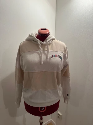 Beige och vit hoodie från Champion - Säljer en beige och vit hoodie från Champion i storlek S. Hoodien har huva med dragsko, broderad Champion-logga på bröstet och liten logga på ärmen. Tillverkad i mjukt bomullsmaterial och har en relaxed fit med breda blockränder.