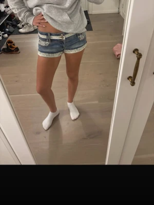 Blå jeansshorts med spetsdetaljer - Säljer ett par blå denimshorts med vita spetsdetaljer vid fickor och benslut. Shortsen har låg midja och klassisk femficksdesign. Perfekta för sommaren och ger en fräsch och trendig look.