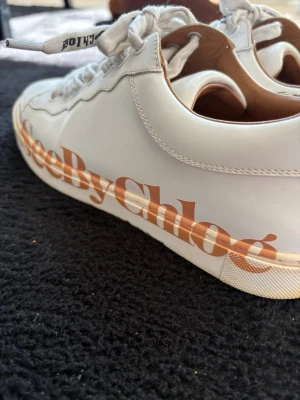 Vita See By Chloé sneakers - Snygga vita sneakers från See By Chloé med stor logga i orange på sidan. Skorna har klassisk låg modell, vita snören och ovandel i slätt läder. Perfekta för dig som vill ha en clean look med en touch av branding. Säljes i använt skick !