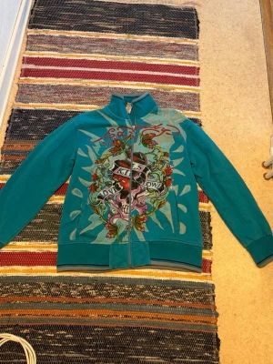 Turkos Ed Hardy Zip-Up med tryck - Turkos Zipup från Ed Hardy med stort färgglatt tryck framtill och logga på ryggen. Använd men i bra skick! 100% bomull