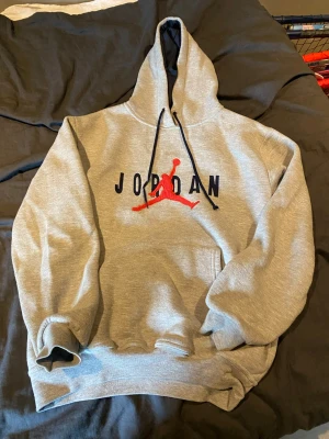 Grå Jordan hoodie med röd logga - Snygg grå hoodie från Jordan med svart text och den klassiska röda Jumpman-loggan på bröstet. Hoodien har huva med svarta snören och en stor magficka. Perfekt för dig som gillar sportig och avslappnad stil.