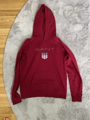Vinröd hoodie från GANT med logga HELT NY ! - Snygg vinröd hoodie från GANT med stor huva och känguruficka. På bröstet finns GANTs logga och ett broderat märke med stjärnor och ränder. Hoodien har långa ärmar och ribbade muddar. Perfekt för en avslappnad och trendig look.