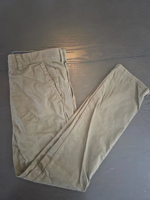 Oliv Gröna chinos från Tommy Hilfiger - Snygga Oliv Gröna chinos från Tommy Hilfiger med klassisk passform och raka ben. Byxorna har sidofickor, bälteshällor och en knappstängning framtill. Tillverkade i mjukt bomullstyg som känns skönt mot huden.