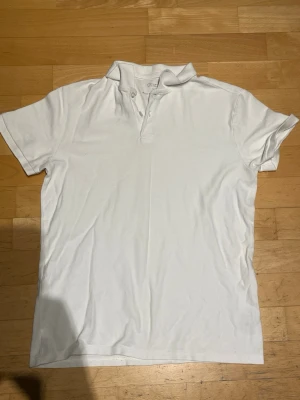 Vit pikétröja med kort ärm - Klassisk vit pikétröja med krage och knappar framtill. Tröjan har korta ärmar och är tillverkad i ett mjukt bomullsmaterial. Enkel och stilren modell som passar till det mesta.