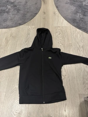 Svart zip-hoodie från Lacoste - Storlek mellan S-MSnygg svart hoodie med dragkedja från Lacoste. Klassisk krokodil-logga på bröstet och två stora fickor framtill. Tillverkad i mjukt material och har en skön huva. Perfekt för en chill och stilren look.
