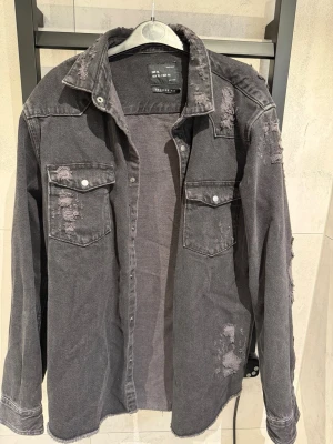Svart distressed jeansjacka - Säljer en svart jeansjacka med slitna detaljer och råa kanter. Jackan har två bröstfickor med lock och silverfärgade knappar. Cool vintagekänsla, perfekt för en avslappnad streetwear-look. Känns som en L.