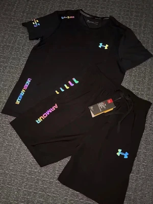 Svart träningsset från Under Armour  - Säljer ett par svart träningsset från Under Armour med färgglada loggor och textdetaljer. Shortsen har snörning i midjan och är tillverkade i ett lätt och sportigt material som passar perfekt för träning eller chill. Snygg och modern design med regnbågsfärgade detaljer. Storlek S, helt ny