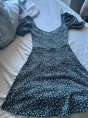 Blommig kortklänning från H&M Divided - Supersöt kortklänning från H&M Divided med svart botten och små vita, gröna och rosa blommor. Klänningen har puffärmar och v-ringning, vilket ger en trendig och feminin look. Perfekt för varma dagar och somriga vibes.
