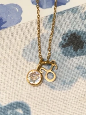 Guld halsband med  - Snyggt halsband som e guldpläterat med ett hänge i form av oxens stjärntecken och ett runt hänge med en glittrande klar sten. Kedjan är tunn och elegant, perfekt för att ge en personlig touch till din stil.