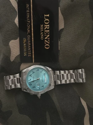Lorenzo Milano silverfärgad klocka - Stilren klocka från Lorenzo Milano med silverfärgad länk i metall och ljusblå urtavla. Urtavlan har arabiska siffror och datumfönster. Boetten har räfflad kant och klockan har en klassisk, elegant look.