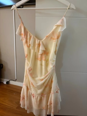 Ljus somrig klänning med volang - Söt kortklänning i ljusa toner med blommigt mönster. Klänningen har tunna axelband, volangdetaljer och omlottkänsla framtill. Perfekt för varma dagar och har en romantisk vibe.