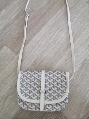 Vit axelväska med grafiskt mönster - Snygg axelväska i vitt med grafiskt mönster i grått och beige. Väskan har justerbar axelrem och stängs med ett lock och magnetknapp. Materialet ser ut att vara konstläder och textil. Perfekt storlek för det viktigaste och riktigt trendig design.