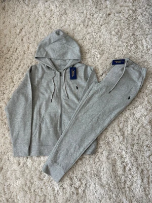 Grått set från U.S. Polo Assn. - Snyggt grått set från U.S. Polo Assn. med hoodie och matchande byxor. Setet har dragkedja på tröjan, fickor och justerbar huva samt resår och dragsko i midjan på byxorna. Båda delarna har den klassiska loggan broderad framtill.