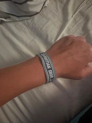 Blått Christian Dior tygarmband - Snyggt blått tygarmband från Christian Dior med texten 'CHRISTIAN DIOR J'ADIOR' broderad i vitt. Armbandet har justerbar knytning med tofsar i ändarna och en stilren, trendig design som passar perfekt till din outfit.