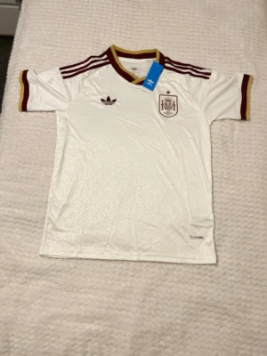 Vit Spanien fotbollströja Adidas - Snygg vit fotbollströja från Adidas med Spaniens landslagsemblem på bröstet. Tröjan har vinröda detaljer på krage, ärmslut och axlar samt Adidas-logga. Tillverkad i lätt och ventilerande material, perfekt för träning eller match.
