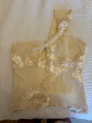 Champagnefärgad oneshoulder topp med spets - Säljer en champagnefärgad oneshoulder topp med stora blommiga spetsdetaljer. Toppen har en unik axelbandslös design på ena sidan och är dekorerad med broderade blommor i samma ton. Perfekt för dig som vill sticka ut med en elegant och trendig look.