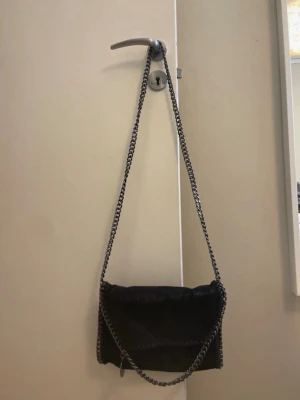 Svart axelväska med kedjerem - Snygg svart axelväska i mjukt material med långa kedjeremmar i silverfärg. Väskan har en enkel, rektangulär form och minimalistisk design som passar till många olika stilar. Perfekt för dig som gillar en stilren look med lite edge.