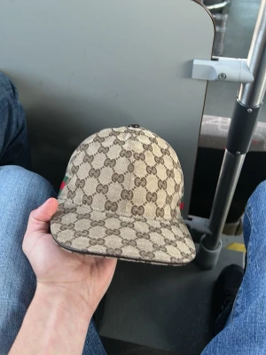 Beige keps med GG-mönster - Säljer en beige keps  med det klassiska GG-monogrammet över hela kepsen. På sidan finns en grön och röd rand som ger extra lyxig känsla. Kepsen har böjd skärm och är tillverkad i canvasmaterial. Perfekt för dig som vill sticka ut med en ikonisk accessoar.
