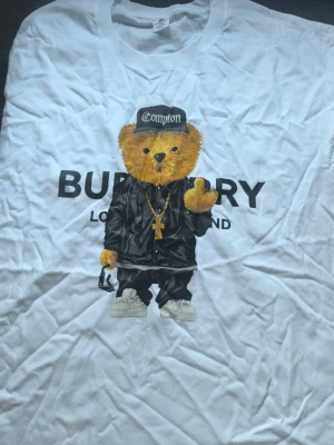 Vit burrbery t shirt med en nalle - Vit t-shirt med stort tryck av en nalle i streetwear-stil, keps och guldkedjor. Texten 'BURBERRY LONDON ENGLAND' syns bakom motivet. Rund halsringning och klassisk passform. Perfekt för dig som vill sticka ut med en unik look.
