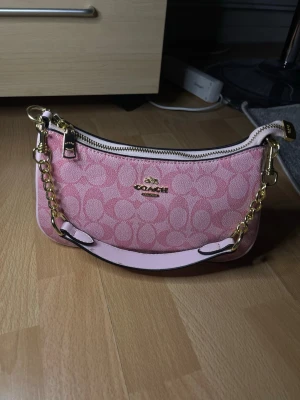 Rosa axelremsväska från Coach - Snygg rosa handväska från Coach med guldiga kedjedetaljer och dragkedja. Väskan har Coachs klassiska mönster i rosa nyanser och en kort axelrem i ljusrosa läder. Perfekt för dig som vill ha en trendig accessoar med exklusiv känsla.