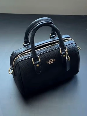 MINI ROWAN BAG  - Coach handväska i svart med dubbla handtag och snygga gulddetaljer. En stilren liten modell med klassisk form, i modellen Mini Rowan Black Bag. All tillbehör medföljer med, i äkta läder.