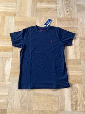 Marinblå t-shirt från Polo Ralph Lauren - Snygg marinblå t-shirt från Polo Ralph Lauren med klassisk röd logga broderad på bröstet. T-shirten har rund halsringning och korta ärmar. Tillverkad i mjuk bomull som känns skön mot huden. Perfekt till jeans eller shorts.