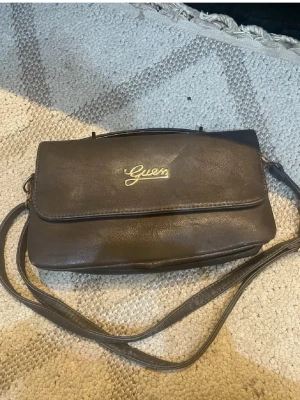 Mörkbrun/grå axelremsväska från Guess - Snygg mörkbrun handväska från Guess i slätt konstläder med guldfärgad logga framtill. Väskan har både axelrem och ett litet handtag i metall, perfekt för dig som vill ha flera bärsätt. Klassisk rektangulär form och stilren design. Svårt att få bild på färgen den är typ grön/grå/brun 
