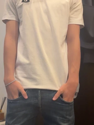 Vit t-shirt med tryck - Säljer en vit t-shirt i bomull med svart tryck på bröstet och ett stort vertikalt tryck på ryggen. Klassisk passform och korta ärmar, perfekt till jeans eller shorts för en clean och enkel stil.
