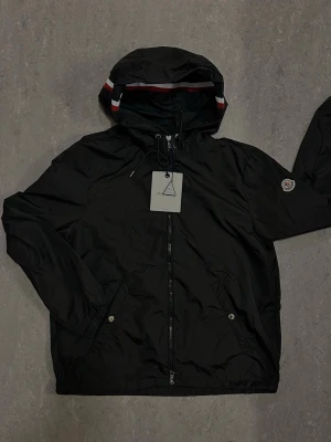 Svart vindjacka från Moncler - Snygg svart vindjacka från Moncler med huva och dragsko. Jackan har dragkedja framtill, två fickor med tryckknappar och Moncler-logga på ärmen. Huvan har röd, vit och blå randig detalj. Perfekt för en sportig och trendig look.