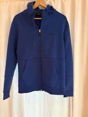 Blå BOSS hoodie med dragkedja och huva - Snygg blå Boss hoodie med dragkedja framtill och huva med dragsko. Hoodien har svarta detaljer på axlarna och stora fickor framtill. Perfekt för en avslappnad och sportig stil. Tillverkad i mjukt material för extra komfort.