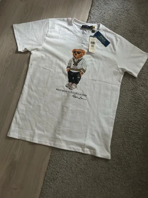Vit Polo Bear t-shirt från Ralph Lauren - Vit t-shirt från Ralph Lauren med Polo Bear-tryck framtill. Klassisk rund hals och korta ärmar. Tillverkad i mjuk bomull för skön känsla. Perfekt för dig som gillar stilrena plagg med en twist.