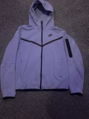 Lila Nike Tech Fleece hoodie - Lila Nike Tech Fleece hoodie med svarta detaljer. Mjuk, bekväm och stilren med hel dragkedja, huva och praktiska fickor. Perfekt för både vardag och träning.