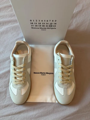 Maison Margiela vita sneakers - Stilrena sneakers från Maison Martin Margiela i vitt och beige med ovandel i läder och mocka. Klassisk låg modell med beige snörning och gummisula. Perfekta för dig som gillar minimalistisk och tidlös design.