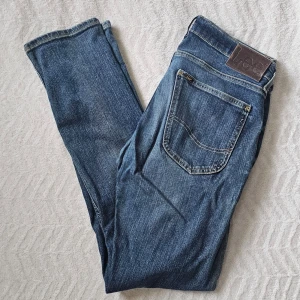 Blå raka jeans från Lee - Säljer ett par klassiska blå jeans från Lee med raka ben och femficksmodell. Jeansen har tydliga kontrastsömmar och Lee-logga på bakfickan samt läderpatch i midjan. Tillverkade i slitstark denim, perfekta till en avslappnad look.