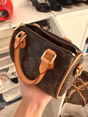 Mini monogram handväska brun - Snygg liten handväska i brunt med klassiskt monogrammönster och detaljer i beige läder. Väskan har två korta handtag och guldfärgade metalldetaljer. Perfekt för dig som gillar trendiga accessoarer och vill ha något unikt till din outfit.
