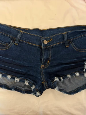 Mörkblå lågmidjade jeansshorts - Säljer ett par mörkblå denimshorts med slitna detaljer och fransig kant. Shortsen har fem fickor, bälteshällor och gul kontrastsöm. Perfekta för sommaren och ger en avslappnad vibe.