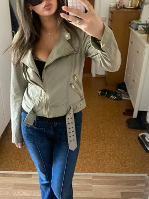Vintage jeansjacka från Vero Moda - Snygg olivgrön jeansjacka från Vero Moda med bälte i midjan och dragkedjor i guld. Jackan har en bikerinspirerad krage och två snedställda fickor framtill. Perfekt att slänga över en t-shirt för en cool look.