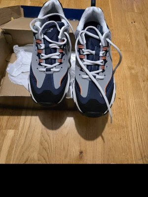 Grå och marinblå sneakers med orange detaljer - Snygga sneakers i grått och marinblått med vita och orange detaljer. Skorna har en chunky sula och vita snören. Perfekta för dig som gillar sportig stil och vill ha något som sticker ut lite extra.