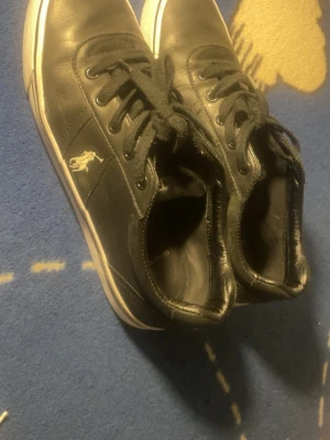 Svarta Polo Ralph Lauren sneakers - Snygga svarta sneakers från Polo Ralph Lauren med klassisk logga på sidan. Skorna har lågt skaft, svarta skosnören och vit sula. Tillverkade i ett slätt material som ger en clean look. Perfekta för dig som gillar stilrena och tidlösa sneakers.