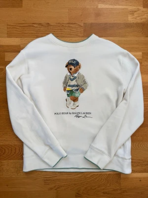 Vit sweatshirt Polo Bear Ralph Lauren storlek L för barn - Vit sweatshirt med ikoniskt Polo Bear-motiv från Ralph Lauren på magen. Tröjan har rund halsringning och ribbade muddar. Trycket visar en nalle med keps. Perfekt för dig som gillar klassisk streetwear med en twist. Storlek L barn (14-16Y). Nypris: 1495kr. 