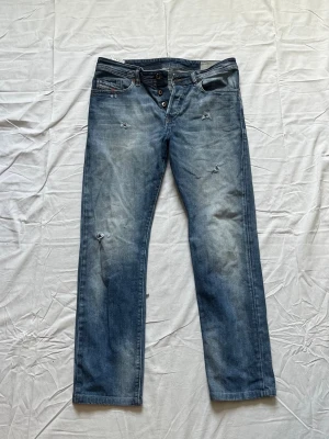 Blå slitna jeans med raka ben - Säljer ett par blå jeans med raka ben och slitna detaljer. Jeansen har knappgylf och klassiska fem fickor. Materialet är denim i bomull och de har en avslappnad passform som passar till det mesta.