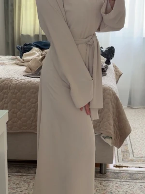 Långärmad maxiklänning med knytband  - En stilren beige långklänning med rund halsringning och trekvartsärmar. Klänningen har en rak passform och är tillverkad i ett mjukt, lätt material som ger ett elegant fall.