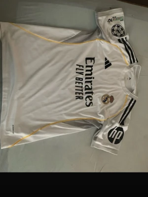 Real Madrid Mbappé matchtröja - Säljer en vit Real Madrid matchtröja från Adidas med Mbappé och nummer 10 på ryggen. Tröjan har svarta detaljer, gula linjer och flera tryck, inklusive klubbmärke och Champions league märke. Köptes för 989 men storleken passar inte mig så därför säljer jag denna varan 