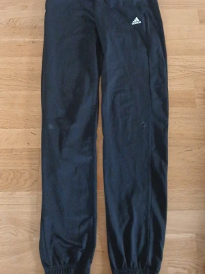 Svarta träningsbyxor från Adidas - Säljer ett par svarta träningsbyxor från Adidas med klassisk logga på vänster lår. Byxorna har resår i midjan och vid bensluten för en bekväm passform. Perfekta för gymmet eller löprundan.