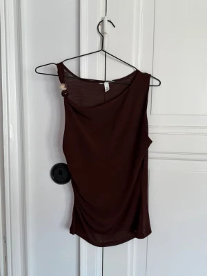 H&M - Brun oneshoulder topp med knappdetalj - Säljer en brun oneshoulder topp med snygg drapering och dekorativ knapp på axeln. Tunn och luftig, perfekt för varma dagar eller som ett statement-plagg. Passar dig som gillar trendiga och unika detaljer.