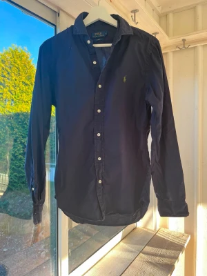 Mörkblå skjorta från Polo Ralph Lauren - Säljer en mörkblå skjorta från Polo Ralph Lauren i slim fit-modell. Skjortan har klassisk krage, vita knappar och det ikoniska broderade logotypen på bröstet. Perfekt för dig som gillar stilrena och tidlösa plagg. Vet ej om den är Dam eller Herr, passar bra på båda! så sätter den som unisex,