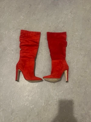 Röda höga stövlar med klack - Snygga röda stövlar i mockaimitation med hög smal klack och spetsig tå. Stövlarna har slouchy skaft och dragkedja på insidan för enkel påtagning. Perfekta för att lyfta vilken outfit som helst.